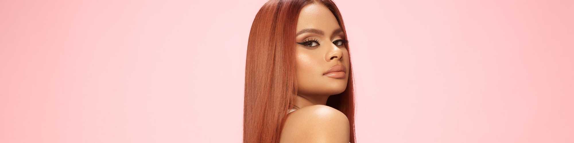 A close up face view of a beautiful woman in an auburn red wig on a pink background.